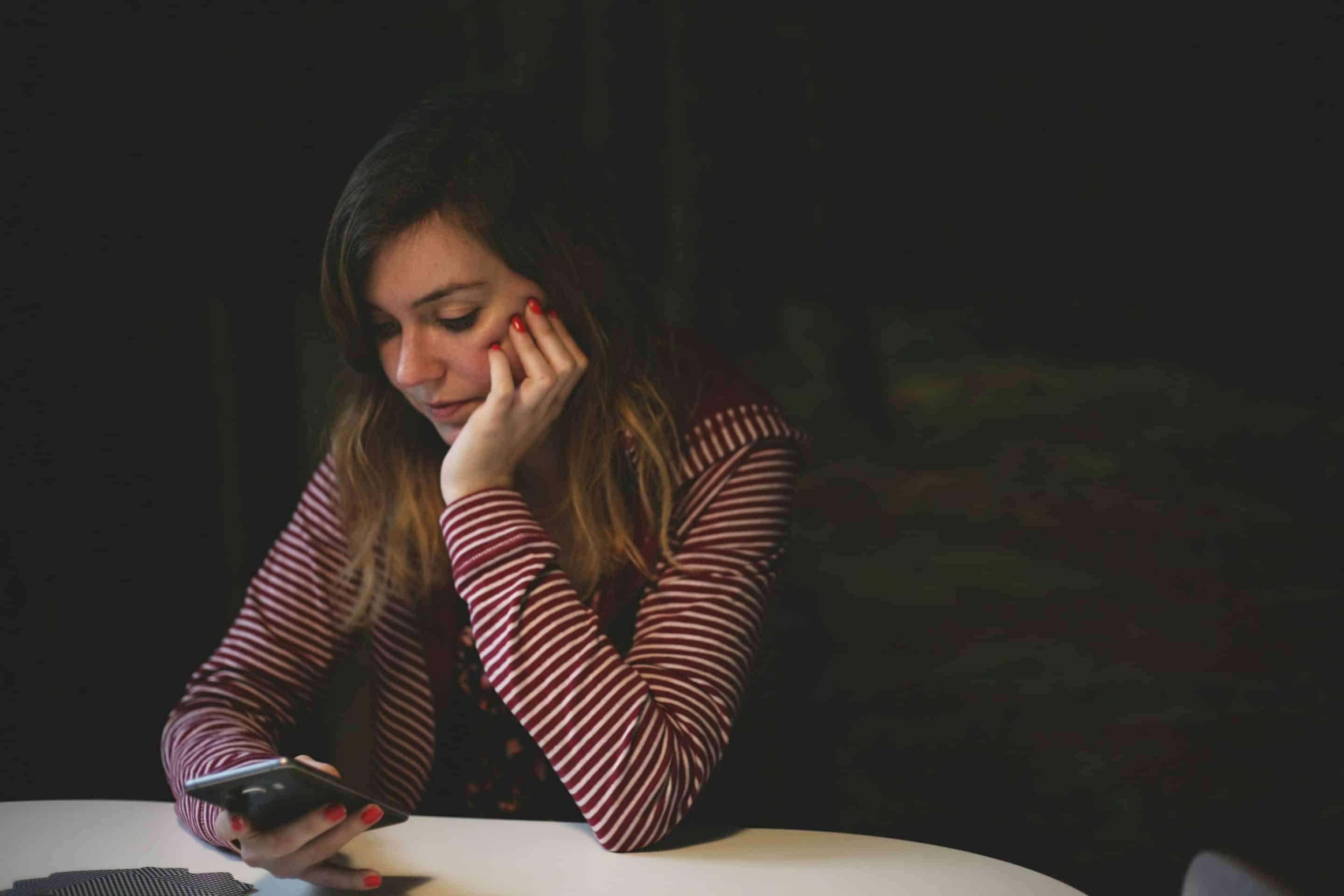 Woman sitting at a table looking at her phone with a worried, thoughtful expression